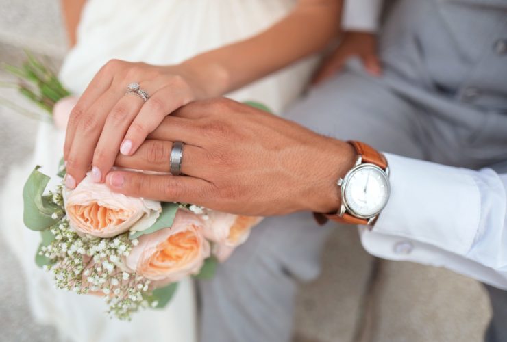 A man and a woman wearing wedding rings hold hands over a bridal bouquet