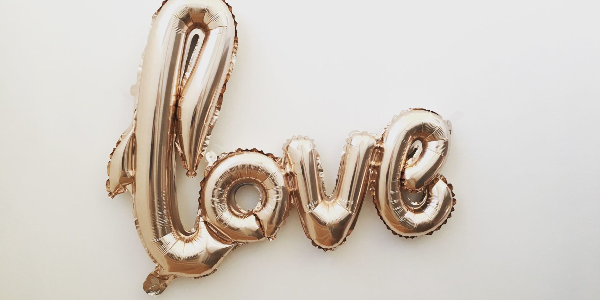 Gold mylar letter balloons that spell "Love" in cursive letters against an off-white color background.