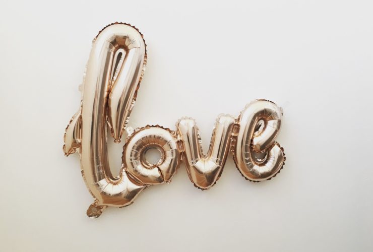 Gold mylar letter balloons that spell "Love" in cursive letters against an off-white color background.
